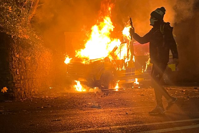 A protester films and a police vehicle burns in the background as a demonstration outside a hotel housing asylum seekers turns violent in Saggart, south-west of Dublin in Ireland on October 21, 2025. Police in riot gear clashed with at least 500 protesters in Dublin on Tuesday outside an asylum seeker hotel, following allegations that a 10-year-old girl was sexually assaulted. (Photo by Peter Murphy/AFP Photo)