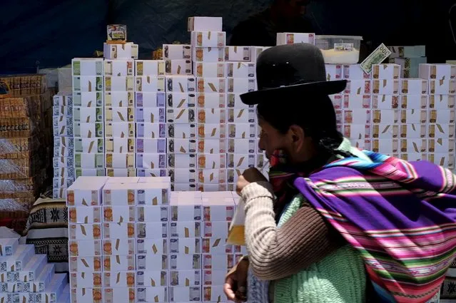 An Aymara woman stands next to a stall displaying miniature bundles of fake money to sell during the “Alasitas” fair in La Paz, Bolivia, January 19, 2016. During the “Alasitas” fair next January 24, Bolivians buy miniature versions of goods they would like to own at the fair, with the hope of acquiring them in real life during the year. (Photo by David Mercado/Reuters)