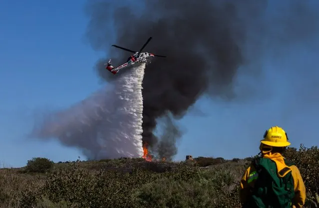 A firefighter watches as a helicopter drops water on a smoldering hillside on February 10, 2022 in Laguna Beach, California. A wind-driven brush fire that broke out in the hills near Laguna Beach amid high temperatures and low humidity forced residents to flee their homes early today. (Photo by Apu Gomes/Getty Images)