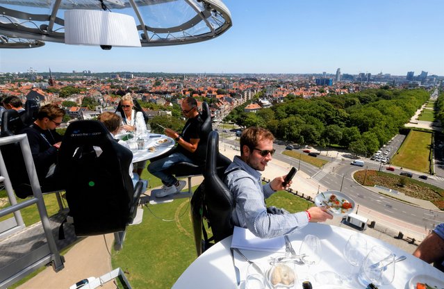 People sit at one of the eight tables on a nine ton structure, suspended from a crane at a height of fifty metres, that can accommodate a total of thirty two guests for a meal during an event known as “Dinner in the Sky” outside Koekelberg Basilica in Brussels, Belgium on June 14, 2022. (Photo by Yves Herman/Reuters)