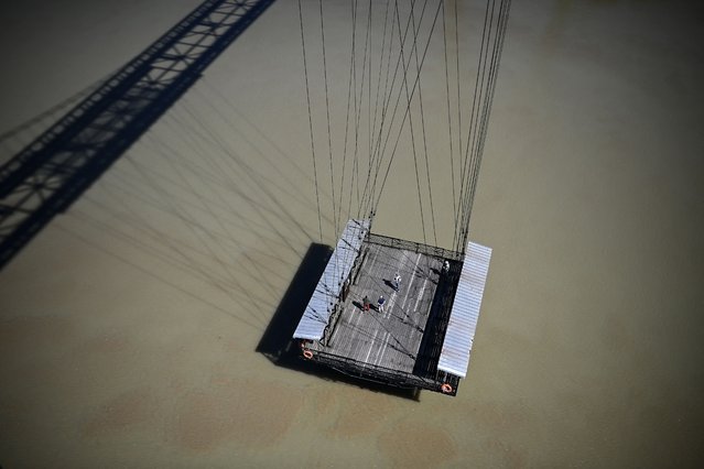Commuters stand on a nacelle suspended from the metal deck of an aerial ferry bridge to cross the Charente river in Rochefort, western France on May 22, 2025. Rochefort's aerial ferry bridge was inaugurated in 1900, allowing people to cross the river without disturbing maritime navigation to and from the city's dockyard. It was used as the main crossing way over the Charente river until 1967, and since 1994 carries pedestrians and cyclists, as the viaduct of the Charente estuary opened nearby in 1991. (Photo by Christophe Archambault/AFP Photo)