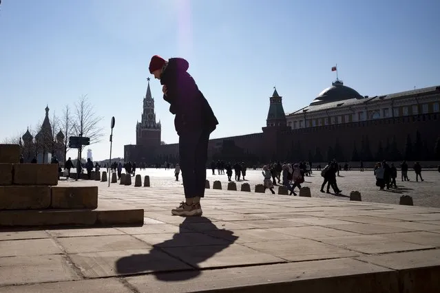 A woman crosses herself in front of Church of Our Lady of Kazan in Red Square with the St. Basil's Cathedral, left, and the Spasskaya Tower, second left, and the Kremlin Wall in the background, in Moscow, Russia, Sunday, March 20, 2022. (Photo by AP Photo/Stringer)