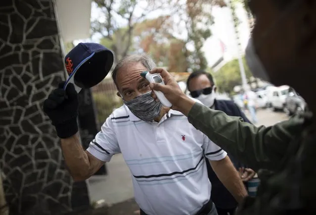 A man removes his cap as a soldier takes his temperature outside a food market as a preventative measure against the spread of the coronavirus in Caracas, Venezuela, Thursday, March 19, 2020. To enter the market, people are required to get their temperature taken and disinfect their hands. (Photo by Ariana Cubillos/AP Photo)