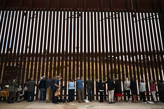 People lay their hands on the border wall and pray towards Mexico during Pastor Seth Clark's last service for The Border Church at the stretch of the US-Mexico border fence known as “Whiskey 8” in San Diego, California on May 25, 2025. As pastor Seth Clark retires from his leadership role with The Border Church the mission of the interdenominational Christian ministry to maintain presence and sacramental solidarity appears more challenging now than ever. While weekly binational communion services started in 2011 on both sides of the US and Mexico border, the US side of the International Friendship Park has remained closed since February 2020 with fears it will never reopen. (Photo by Patrick T. Fallon/AFP Photo)