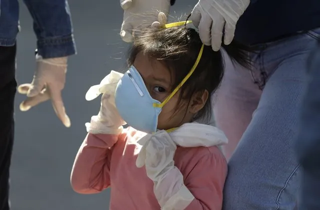 A girl wearing plastic gloves is helped with her mask before traveling with her family to her home province of Piura, at the bus station in Lima, Peru, Monday, April 27, 2020. After a week of not being allowed to leave the capital under strict quarantine rules to help contain the spread of the new coronavirus, day laborers and informal workers and their families are now allowed to travel home, but local authorities are screening them for COVID-19 before letting them travel. (Photo by Martin Mejia/AP Photo)