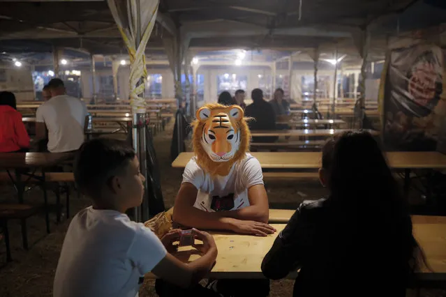 In this picture taken Thursday, September 12, 2019, children, one wearing a tiger mask, sit at a table in the food area of an autumn fair in Titu, southern Romania. Romania's autumn fairs are a loud and colorful reminder that summer has come to an end and, for many families in poorer areas of the country, one of the few affordable public entertainment events of the year. (Photo by Vadim Ghirda/AP Photo)