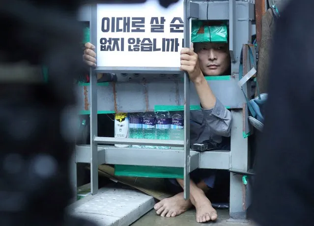 Yoo Choi-ahn, vice chief of the subcontractor union, stuck inside a steel cage-like structure that he welded himself on the floor of the occupied oil tanker holds a sign during a strike at Daewoo Shipbuilding & Marine Engineering in Geoje, South Korea, July 19, 2022. The sign reads “We can't live like this”. (Photo by Yonhap via Reuters)