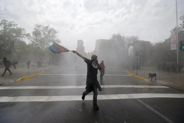 A Mapuche Indian activist waves a Mapuche flag as he covers his face as riot police release water laced with tear gas during a protest march by Mapuche Indian activists against Columbus Day in downtown Santiago, Chile, October 12, 2015. (Photo by Ivan Alvarado/Reuters)