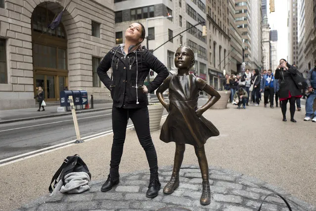 A woman strikes a pose in front of a statue titled “Fearless Girl” on Wednesday, March 8, 2017, in New York. A big investment firm, State Street Global Advisors, put the statue there to highlight International Women's Day. The work by artist Kristen Visbal. (Photo by Mark Lennihan/AP Photo)