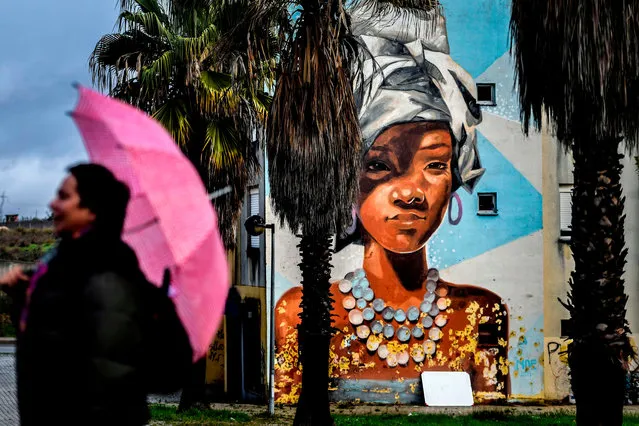 A visitor holds her umbrella close to a mural by Uruguayan artist Maria Noe during a guided visit to Quinta do Mocho neighbourhood in Sacavem, outskirts of Lisbon, on November 11, 2019. The image of a black woman removing her white-woman mask is one of the hundreds of murals that cover the buildings of the underprivileged district of Quinta de Mocho in Lisbon, that has been transformed by street art. (Photo by Patricia De Melo Moreira/AFP Photo)
