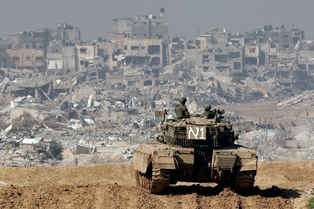 A picture taken from a position in southern Israel along the border with the Gaza Strip on January 19, 2024, shows an Israeli tank rolling along the fence as damaged buildings are see in the Gaza strip amid continuing battles between Israel and the militant group Hamas. (Photo by Jack Guez/AFP Photo)