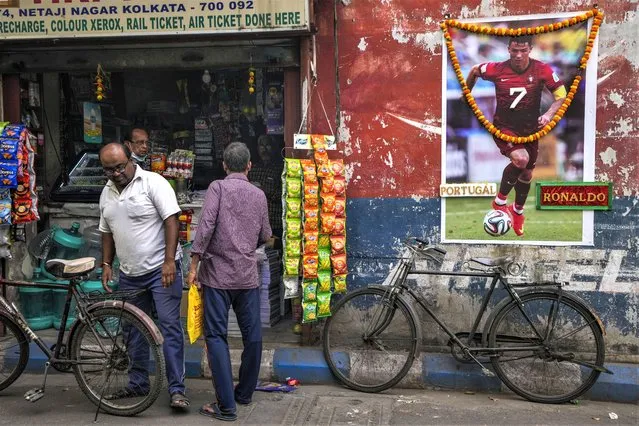 People buy grocery from a shop next to a garlanded photograph of Portugal's Cristiano Ronaldo put up by fans to mark the ongoing soccer World Cup in Kolkata, India, Thursday, December 8, 2022. (Photo by Bikas Das/AP Photo)