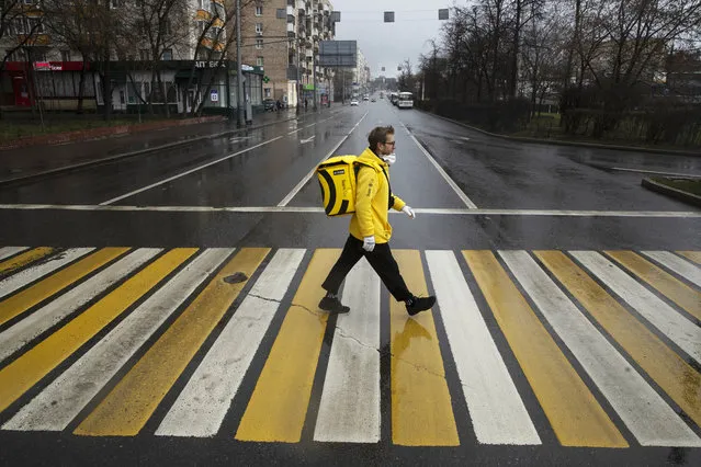 In this photo taken on Thursday, April 16, 2020, Russian businessman Sergey Nochovnyy wearing a face mask to protect against coronavirus crosses a road, on his way to pick up a food order to deliver, in Moscow, Russia. A Russian businessman has become a delivery man amid the nation’s partial economic shutdown due to the coronavirus. Nochovnyy said that he hasn’t lost his business and made the move to “change an angle” and avoid being locked at home amid the epidemic. (Photo by Pavel Golovkin/AP Photo)