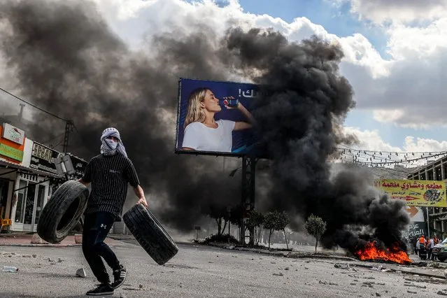 A masked man walks with tires past other burning tires during clashes at the village of Deir Sharaf near the western entrance of the city of Nablus in the occupied West Bank on October 20, 2022, as Palestinian protesters attempt to remove a barrier erected by Israeli forces to lock down Nablus. (Photo by Jaafar Ashtiyeh/AFP Photo)