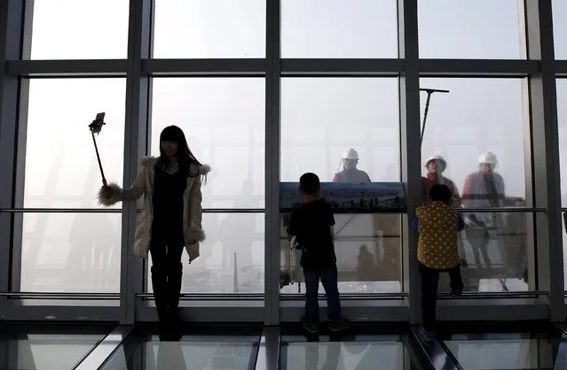 A woman uses a selfie stick to take a picture while workers clean the exterior of the Shanghai World Financial Center skyscraper amid heavy smog at the financial district of Pudong in Shanghai, China, December 25, 2015. (Photo by Aly Song/Reuters)