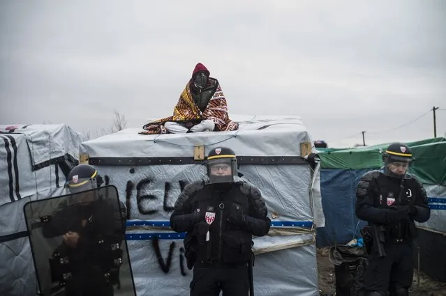 A migrant stand on his makeshift shelter as police secure the Jungle migrant camp during its demolition in Calais, France, 01 March 2016. A court in Lille, northern France, upheld an expulsion order issued by local authorities to clear part of the migrant camps. Tensions rose on 29 February 2016 as riot police fired tear gas in the direction of onlookers while workers bulldozed the makeshift huts. (Photo by Yoan Valat/EPA)