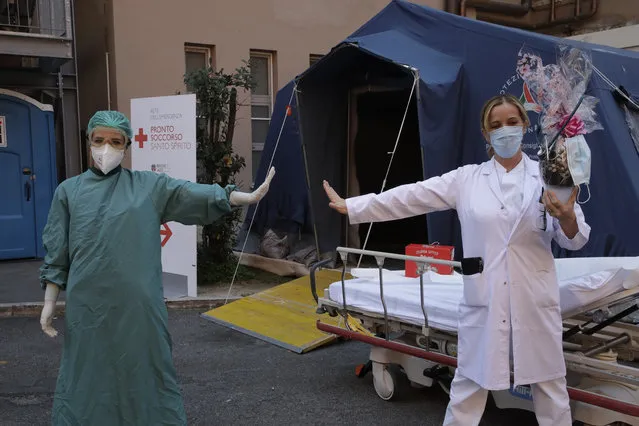 Monica Carfora, right, deputy head of the emergency of the Santo Spirito Hospital, which now has a separate emergency for possible covid patients, holds a chocolate Easter egg as she poses for a photograph with nurse Silvia Sforza, holding out their arms to indicating social distancing, outside the entrance to for covid emergency, in central Rome, Sunday, April, 12, 2020. (Photo by Alessandra Tarantino/AP Photo)