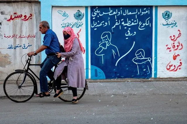 Syrians ride a bicycle past a mural painted as part of an awareness campaign by the United Nations International Children's Emergency Fund (UNICEF) and World Health Organization (WHO) intitative, bearing instructions on protection from COVID-19 in the Kurdish-majority city of Qamishli of Syria's northeastern Hasakeh province on August 16, 2020, after a spike in infections in the area. (Photo by Delil Souleiman/AFP Photo)