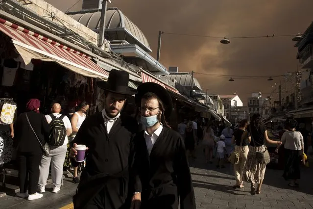 Ultra-Orthodox Jews stroll through the Machane Yehuda market under a sky darkened by nearby wildfires, in Jerusalem, Sunday, August 15, 2021. A massive wildfire outside of Jerusalem on Sunday sent a thick cloud of smoke over the city as authorities struggled to contain the blaze. (Photo by Maya Alleruzzo/AP Photo)