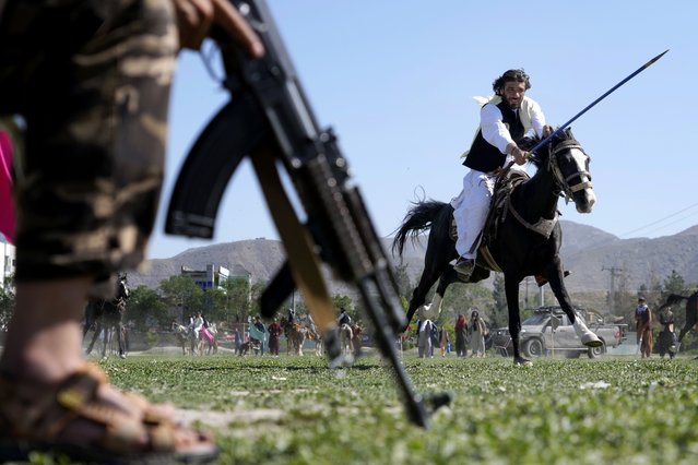 An Afghan man rushes to the target with his horse as a Taliban fighter stands guard during a spear racing in the sprawling Chaman-e-Huzori park in downtown Kabul, Afghanistan, Friday, May 6, 2022. (Photo by Ebrahim Noroozi/AP Photo)