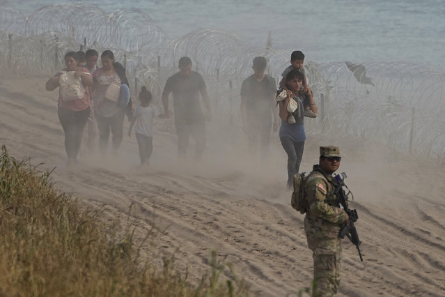 A Texas guardsman watches as migrants who crossed the Rio Grande from Mexico to the U.S. walk along concertina wire, Monday, July 31, 2023, in Eagle Pass, Texas. (Photo by Eric Gay/AP Photo)