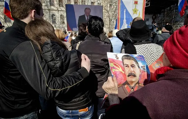 An elderly woman holds a calendar depicting former Soviet leader Josef Stalin while watching a broadcast of Russian President Vladimir Putin's speech on Crimea, seen in background, in Sevastopol, Crimea, Tuesday, March 18, 2014, as thousands of pro-Russian people gathered to watch the address.  Fiercely defending Russia's move to annex Crimea Putin said Russia had to respond to what he described as a western plot to take Ukraine into its influence. (Photo by Vadim Ghirda/AP Photo)