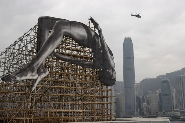 A helicopter flies near the art piece “GIANTS: Rising Up” at Harbour City in Hong Kong China, 13 March 2023. The 12-meter-high and 12-meter-wide installation by French artist JR depicts a larger-than-life high jumper floating in mid-air adjacent to Victoria Harbour. It will be displayed until 23 April. (Photo by Jerome Favre/EPA)