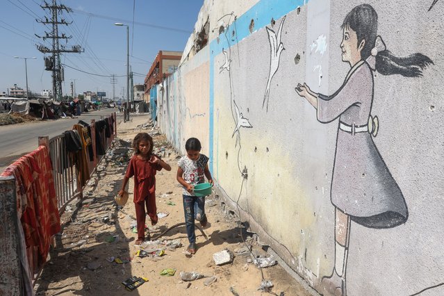 Palestinian girls whalk past a mural at a makeshift displacement camp set up on a roadside in Deir el-Balah in the central Gaza Strip on August 13, 2024 amid the ongoing conflict between Israel and the Palestinian Hamas movement. (Photo by Eyad Baba/AFP Photo)