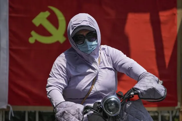 A woman wearing a sun protection clothing rides on an electric bike passes by a communist party flag outside a community in Beijing, Thursday, July 21, 2022. (Photo by Andy Wong/AP Photo)
