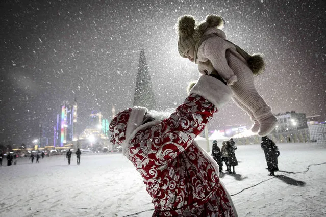 An actor dressed as Ded Moroz (Santa Claus) or Father Frost, plays with a child during snowfall at a square with a Christmas tree and and the main mosque during New Year's celebrations in Grozny, Russia, Wednesday, December 22, 2021. (Photo by Musa Sadulayev/AP Photo)