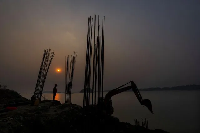 A workers watches an earthmover machine engaged in a construction of a bridge along the river Brahmaputra in Guwahati, India, Tuesday, March 14, 2023. India’s annual budget of $550 billion this year calls for ramping up capital spending by 33% to spur economic growth and create jobs ahead of a general election next year. (Photo by Anupam Nath/AP Photo)