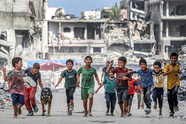 Children walk with a dog past destroyed buildings along a street in Khan Yunis in the southern Gaza Strip on July 8, 2024 amid the ongoing conflict in the Palestinian territory between Israel and Hamas. (Photo by Bashar Taleb/AFP Photo)