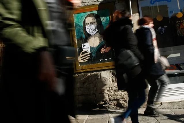 People walk by a poster by Italian urban artist Salvatore Benintende aka “TVBOY” depecting Leonardo da Vinci's Mona Lisa  wearing a protective facemask and holding a mobile phone reading “Mobile World Virus” in a street of Barcelona on February 18, 2020, a week after the World Mobile Congress was cancelled due to fears stemming from the coronavirus that sparked an exodus of industry heavyweights. (Photo by Pau Barrena/AFP Photo)