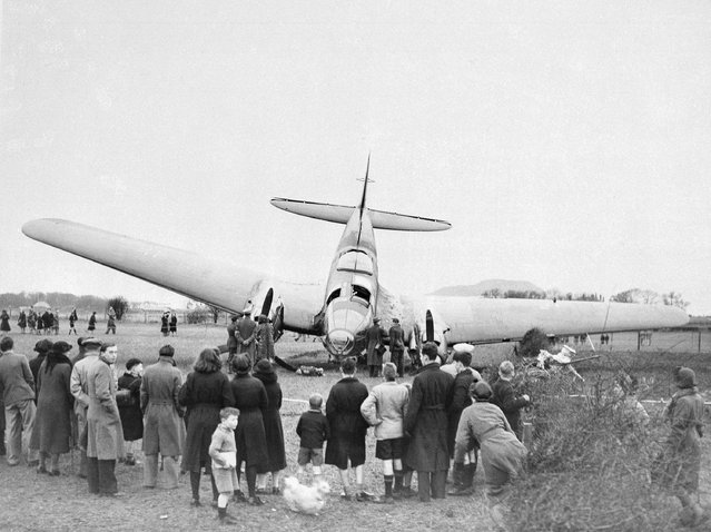Crowds of curious English people are shown gathered around this German bomber, March 8, 1940, after it was shot down, somewhere in England, by an R.A.F., fighter. The Nazi plane crashed after narrowly missing some telegraph wires, ending up with its tail in the air. (Photo by AP Photo)
