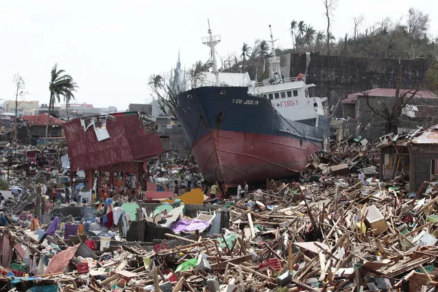 Survivors walk past a ship that lies on top of damaged homes after it was washed ashore in Tacloban city, Leyte province central Philippines on Sunday, November 10, 2013. The city remains littered with debris from damaged homes as many complain of shortage of food, water and no electricity since the Typhoon Haiyan slammed into their province. Haiyan, one of the most powerful typhoons ever recorded, slammed into central Philippine provinces Friday leaving a wide swath of destruction and scores of people dead. (Photo by Aaron Favila/AP Photo)