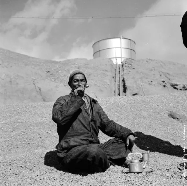 1954:  An Iranian oil worker in an arid landscape, an oil tank in the distance, sits enjoying the labourer's staple diet of bread and tea