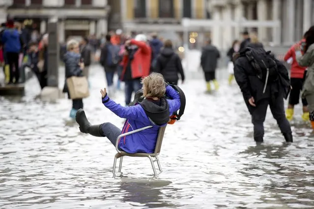 A woman sits as tourists make their way through flooded St. Mark's Square in Venice, Italy, Thursday, November 1, 2018 as rainstorms and strong winds have been battering the country. Two people were killed when a falling tree crushed their car in the mountainous countryside in northwestern Italy, as rainstorms and strong winds continued to pummel much of the country. (Photo by Luca Bruno/AP Photo)