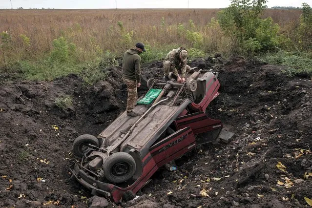 Ukrainian soldiers scavenge from a destroyed car near Prudyanka, Kharkiv region, on September 26, 2022, amid the Russian invasion of Ukraine. Ukraine's latest suspected mass burial site is in a shell-damaged and abandoned industrial chicken farm, on a hill near the Russian border strewn with the debris of battle. It is not known how many bodies lie there – troops and officials speak of 90 to 100 without saying how they know – but the signs of recent violence lie in the rubble all around. (Photo by Yasuyoshi Chiba/AFP Photo)