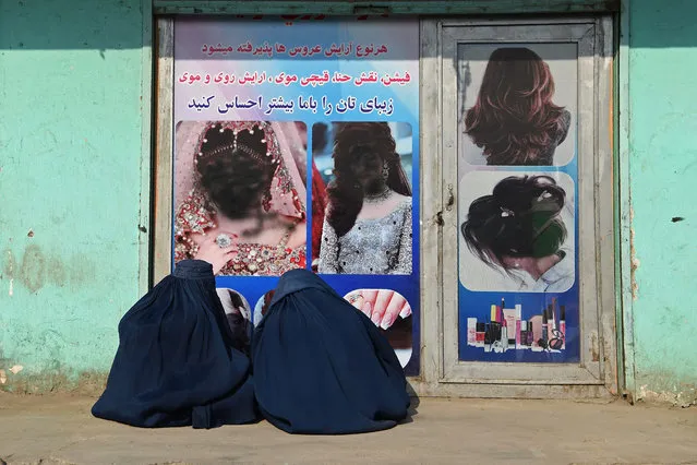 Afghan burqa-clad women sit in front of a beauty salon with images of women defaced using spray paint in Jalalabad on December 13, 2021. (Photo by Wakil Kohsar/AFP Photo)