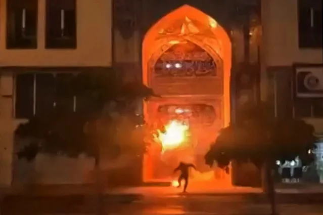 This image grab from a UGC video posted on December 11, 2022, reportedly shows a protester placing an object at the entrance of the Khomeini Seminary in Iran's Bushehr city, before fleeing the scene as the object erupts in flames. (Photo by UGC/AFP Photo)