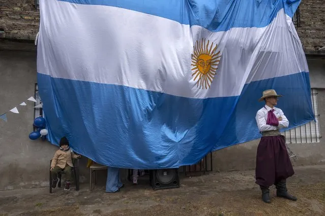 A child sits next to a giant national flag as a neighbor dressed as a gaucho waits to perform in a folkloric dance during the Independence Day celebrations in La Matanza, on the outskirts of Buenos Aires, Argentina, Wednesday, May 25, 2022. (Photo by Rodrigo Abd/AP Photo)