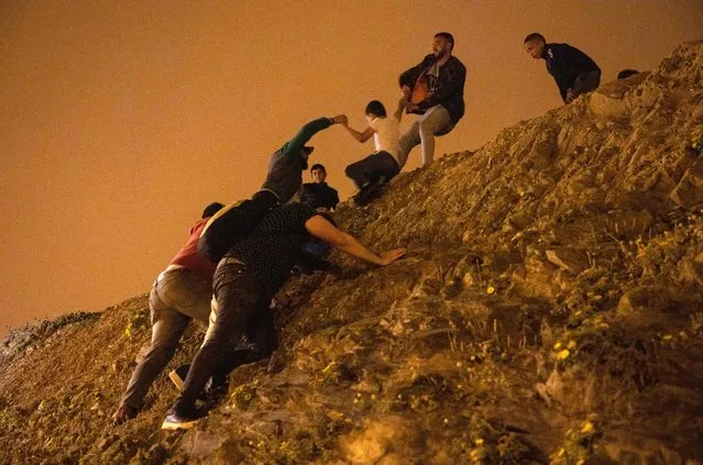 Moroccan migrants climb a rocky cliffside as a policeman watches in the northern town of Fnideq as they attempt to cross the border from Morocco to Spain's North African enclave of Ceuta on May 18, 2021. At least 5,000 migrants, an unprecedented influx at a time of high tension between Madrid and Rabat, slipped into Ceuta on May 17, a record for a single day, Spanish authorities said. They reached the enclave by swimming or by walking at low tide from beaches a few kilometres to the south, some using inflatable swimming rings and rubber dinghies. (Photo by Fadel Senna/AFP Photo)