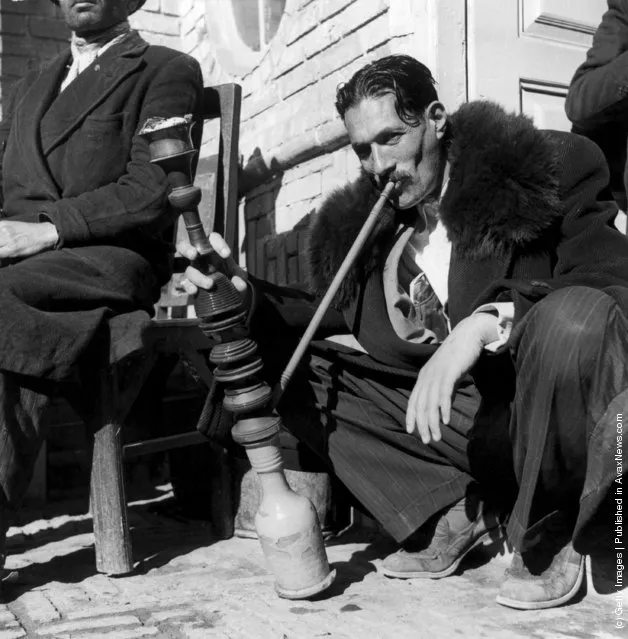 1955:  An Iranian smokes a Persian hookah or  water pipe on the pavement in Isafahan. The pipe is hired by a specialist merchant for single use