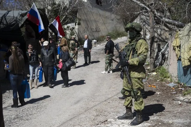 A Russian soldier keeps watch as local residents receive humanitarian aid in the Syrian village of Ghunaymiyah, about 15 kilometers from Turkish border, Tuesday, March 1, 2016. Residents who recently returned to their homes after the government last month captured the village from Nusra Front fighters. (Photo by Pavel Golovkin/AP Photo)