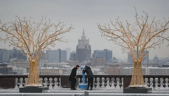 Two men between an artificial trees decorated with light bulbs look at the Moscow buildings through a telescope at the observation deck of Vorobyovy Gory in Moscow, Russia, 08 February 2022. (Photo by Yuri Kochetkov/EPA/EFE)