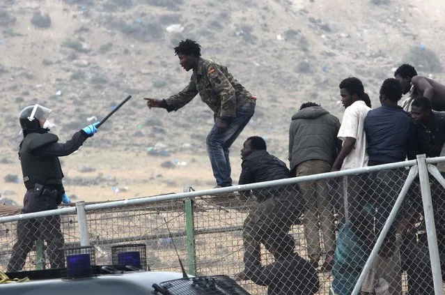 A sub-Saharan migrant agues with a Spanish Guardia Civil officer holding a baton, as they sit on top of a metallic fence that divides Morocco and the Spanish enclave of Melilla, Thursday, May 1, 2014. Spain says around 700 African migrants have rushed its barbed wire border fences in the North African enclave of Melilla, and although police repelled most, 140 managed to enter Spanish territory. (Photo by Fernando Garcia/AP Photo)