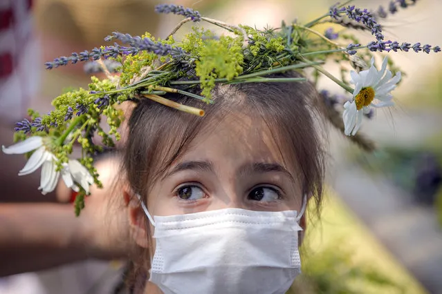 A girl wearing and a face mask to combat the spread of the COVID-19 infections and a flower crown she made herself at a workshop for children stands during an event inspired by pre-Christian traditions at the Dimitrie Gusti Village Museum in Bucharest, Romania, Wednesday, June 24, 2020. According to pre-Christian traditions, fairies, called in Romanian “Sanziene”, come to earth around the summer solstice bringing fertility to land and beings for the coming summer. (Photo by Vadim Ghirda/AP Photo)