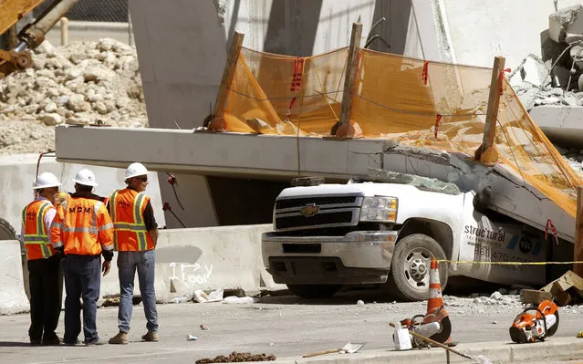 Workers stand next to a section of a collapsed pedestrian bridge, Friday, March 16, 2018 near Florida International University in the Miami area. The new pedestrian bridge that was under construction collapsed onto a busy Miami highway Thursday afternoon, crushing vehicles beneath massive slabs of concrete and steel, killing and injuring several people, authorities said. (Photo by Wilfredo Lee/AP Photo)