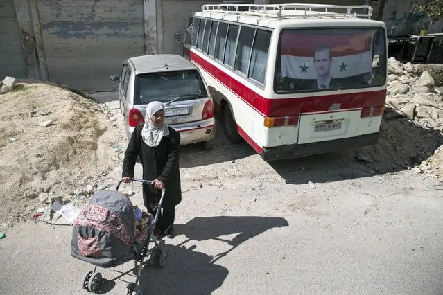 A woman with baby stroller stays near a bus with a portrait of Syrian President Bashar Assad in the window on a road near Latakia in Syria on Wednesday, March 2, 2016. (Photo by Pavel Golovkin/AP Photo)
