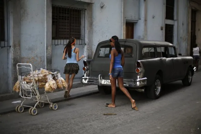 Young women walk in between a shopping cart with baked goods for sale and a vintage car, on a street in Havana December 17, 2014. (Photo by Enrique De La Osa/Reuters)